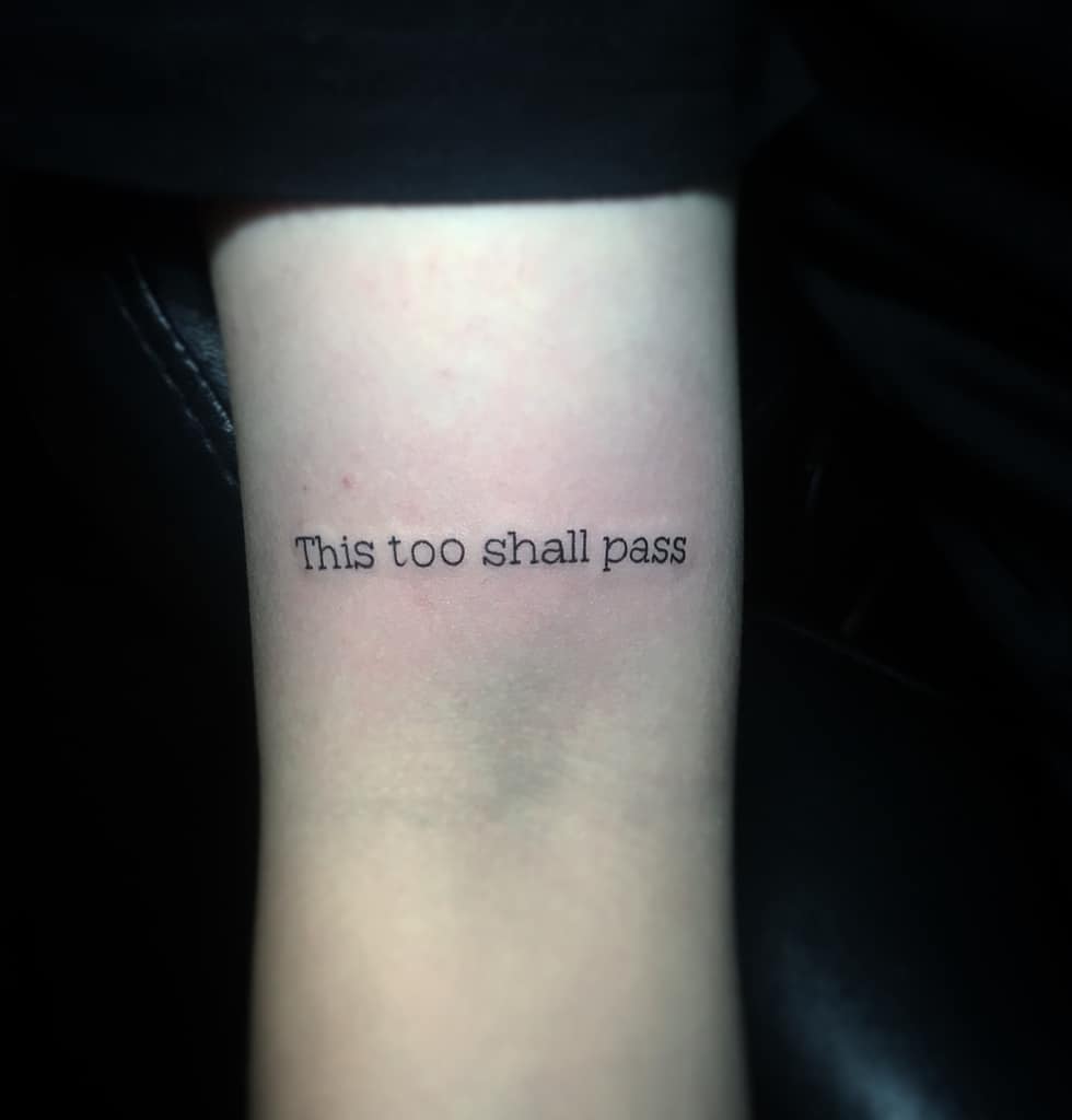 This image shows a tattoo on the arm with the phrase "This too shall pass" in a simple, typewriter-style font.