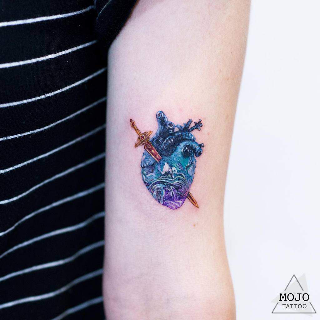 Tattoo of ocean waves and ship inside a heart with a sword through it on an upper arm