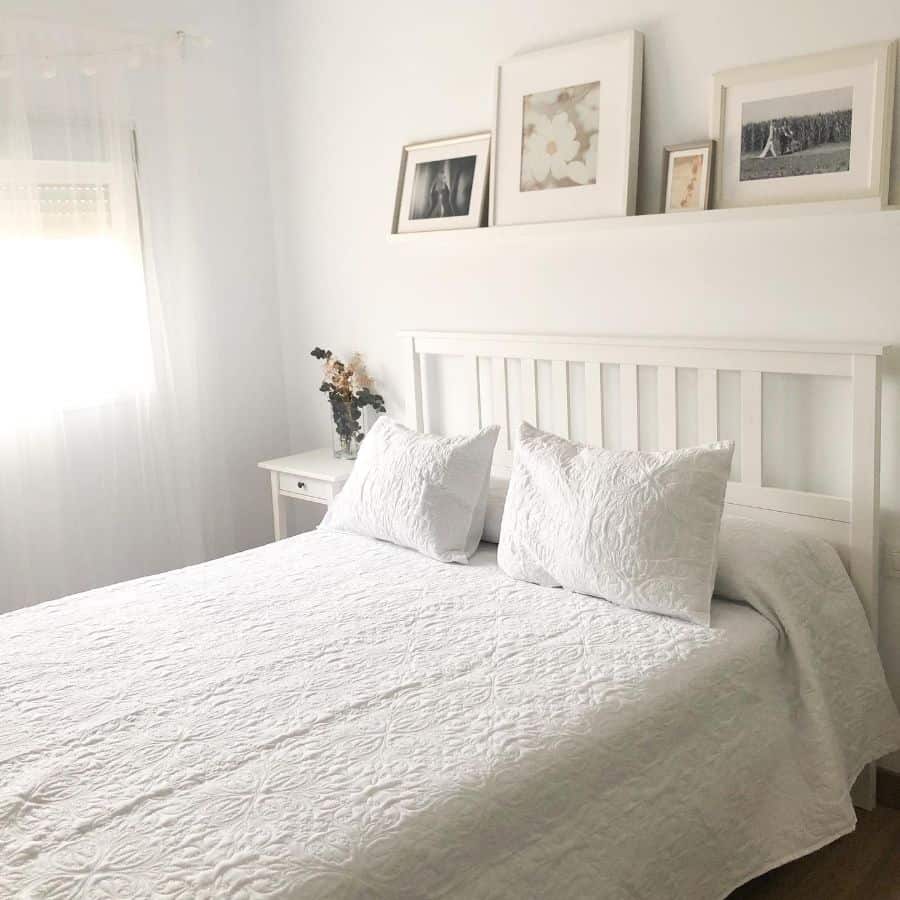 Minimalist bedroom with a white bedspread, framed photos above the headboard, and a small nightstand with flowers