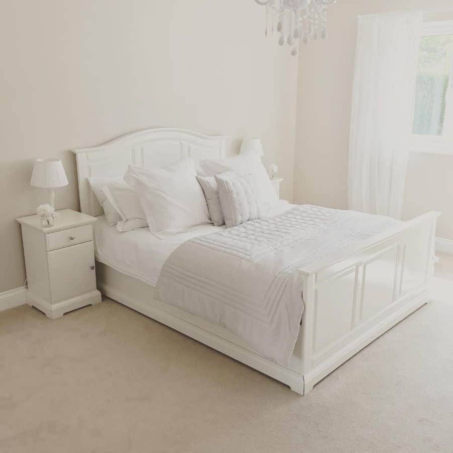 A bright, elegant bedroom with a white bed, matching nightstand, and soft lighting from a window