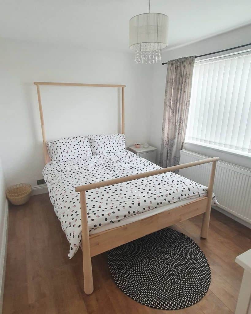 Minimalist bedroom with a wooden bed, polka dot bedding, round rug, white walls, and window with blinds and curtains