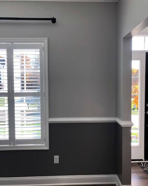 A well-lit room with chair rail trim, a window with white shutters, and a door showing autumn leaves