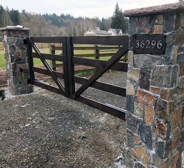 Rustic wooden driveway gate featuring a cross-brace pattern, framed by stone pillars with a house number plaque.