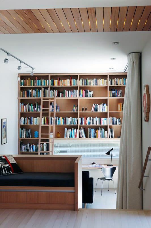 Modern home library with a wall of bookshelves, desk, chair, ladder, and a cozy reading nook with a black cushion