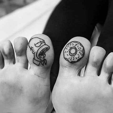 A pair of black ink tattoos on the big toes, one featuring a cartoon character drooling and the other a sprinkled donut, creating a humorous design.