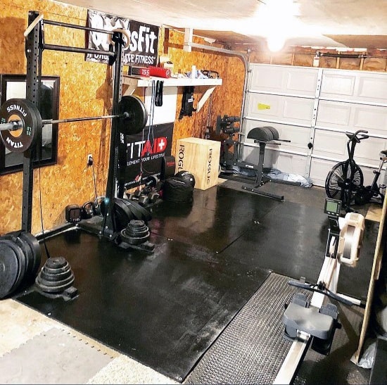 Compact garage gym with squat rack, rower, air bike, weight plates, and GHD machine on rubber flooring.