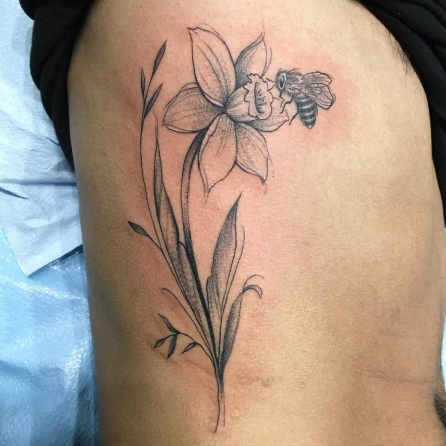 This tattoo showcases a delicate daffodil flower paired with a bee in flight. The thin line work creates a soft, natural feel, while the shading adds depth and realism. The positioning along the arm gives the design a sense of movement, enhanced by the bee's dynamic placement near the flower. The contrast between the floral details and the bee creates a balanced, nature-inspired tattoo.