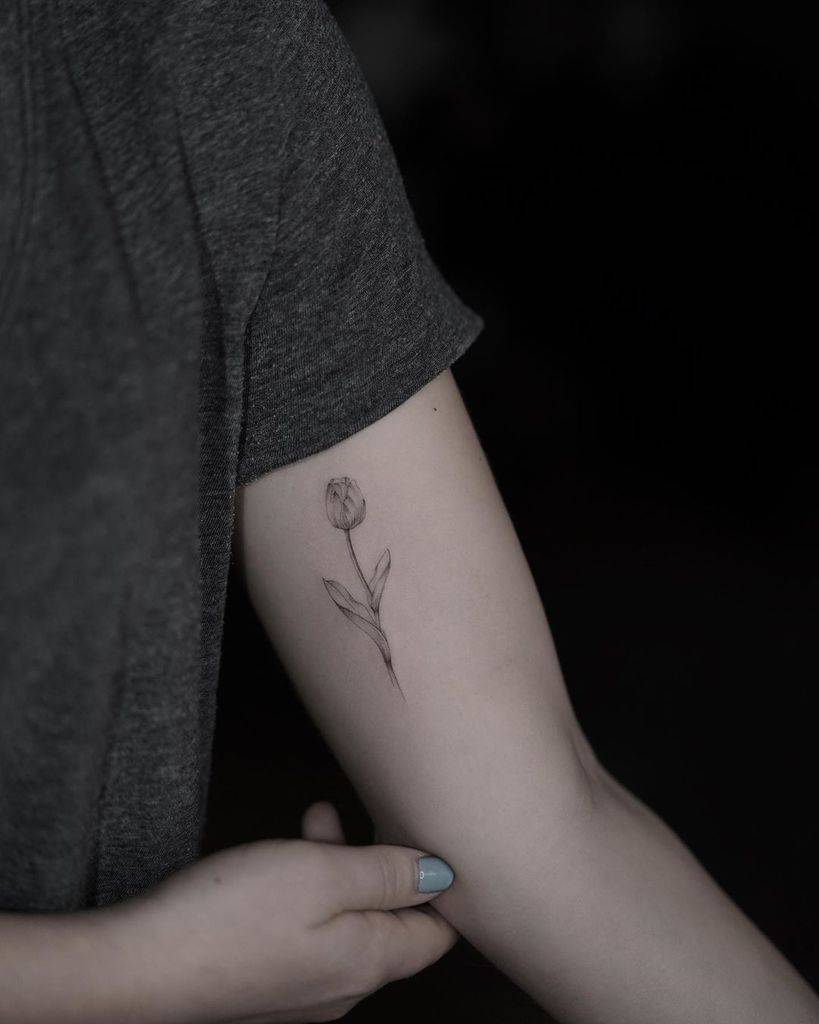 A minimalist tulip tattoo on the upper arm, featuring a simple stem and delicate flower outlined in fine lines.