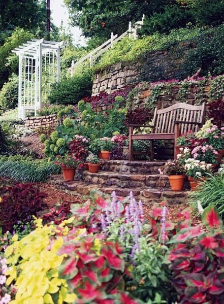 Charming sloped garden with colorful flowers, stone steps, a wooden bench seating area, and a white arbor for a cozy, inviting look