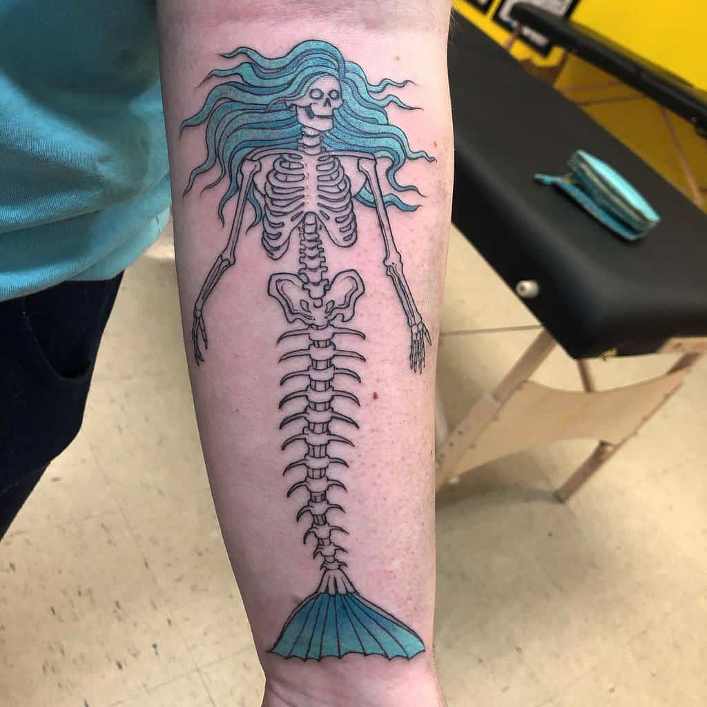 Tattoo of a skeleton mermaid with flowing blue hair, a ribcage upper body, and a detailed spine transitioning into a fishtail.