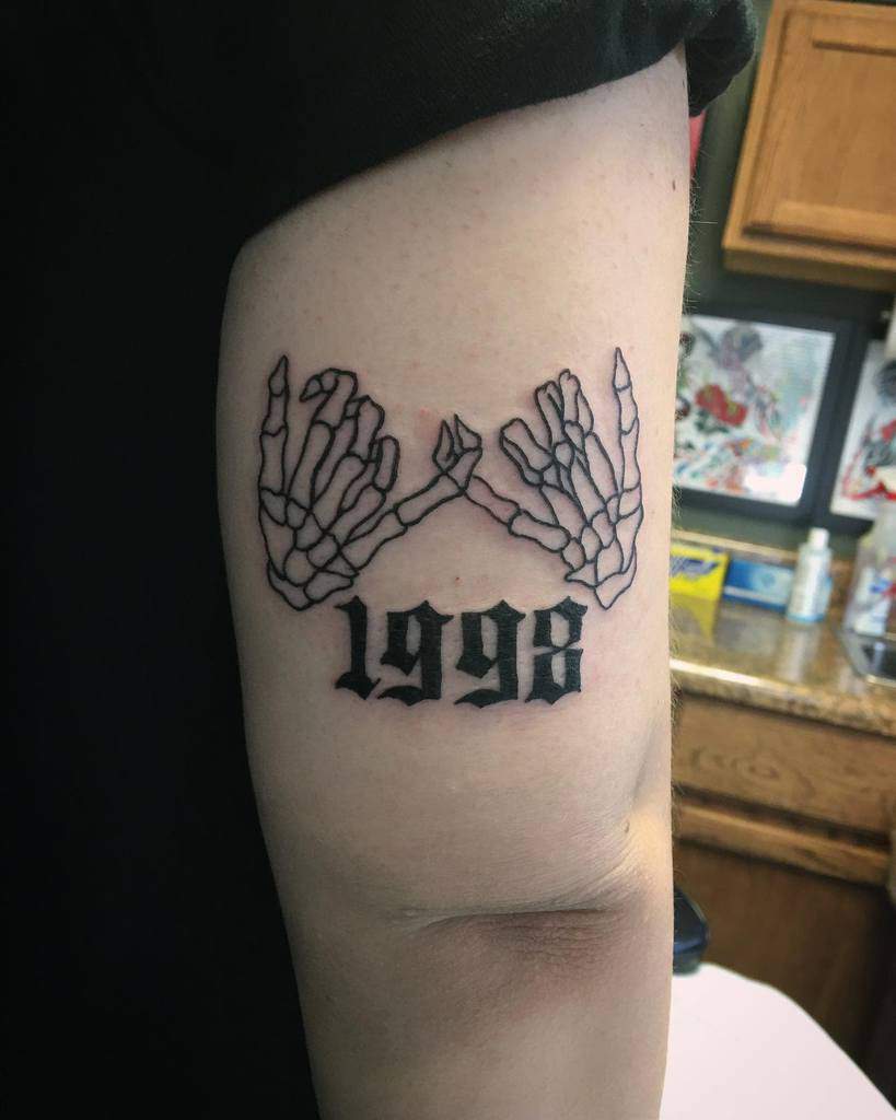 Skeleton pinky promise tattoo with bold line art and the year "1998" in gothic font, placed on the upper arm