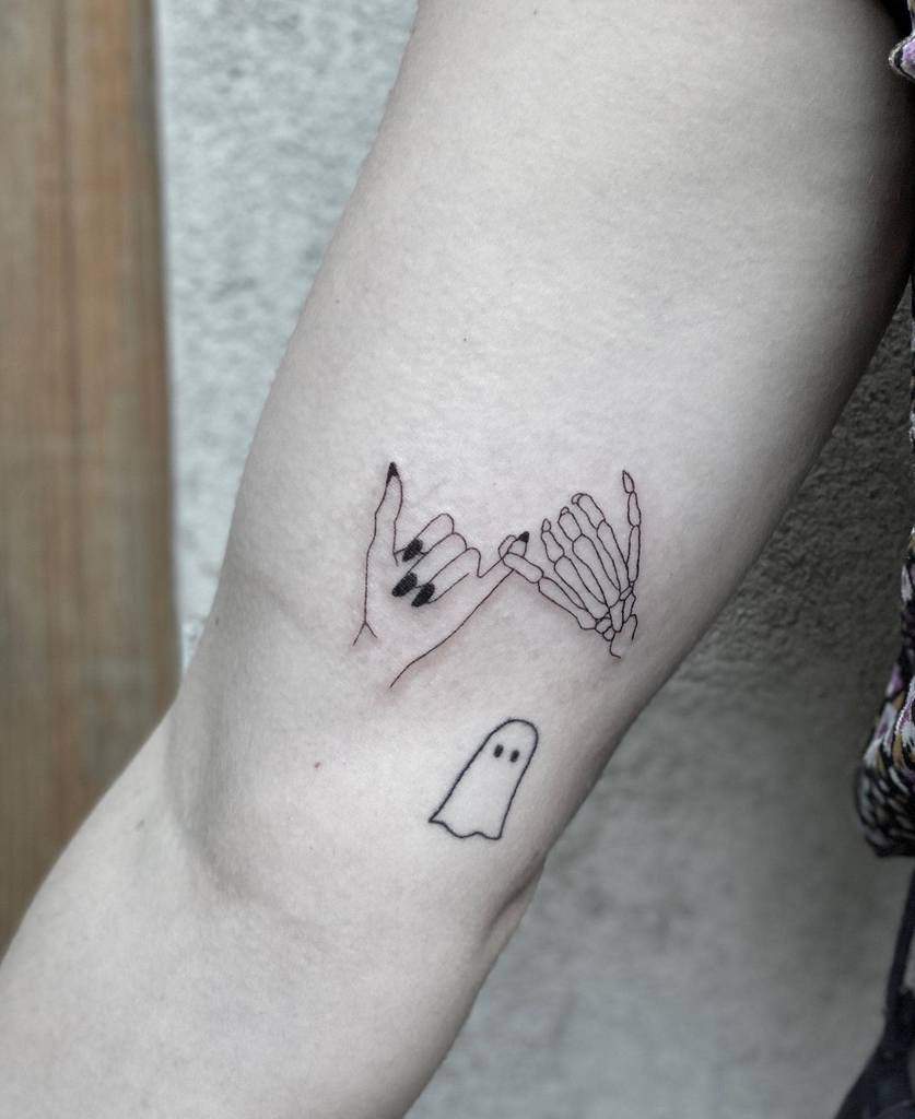 Tattoo of a ghost and interlocking hands on an arm; one hand is skeletal, the other is not, both with pinky fingers linked
