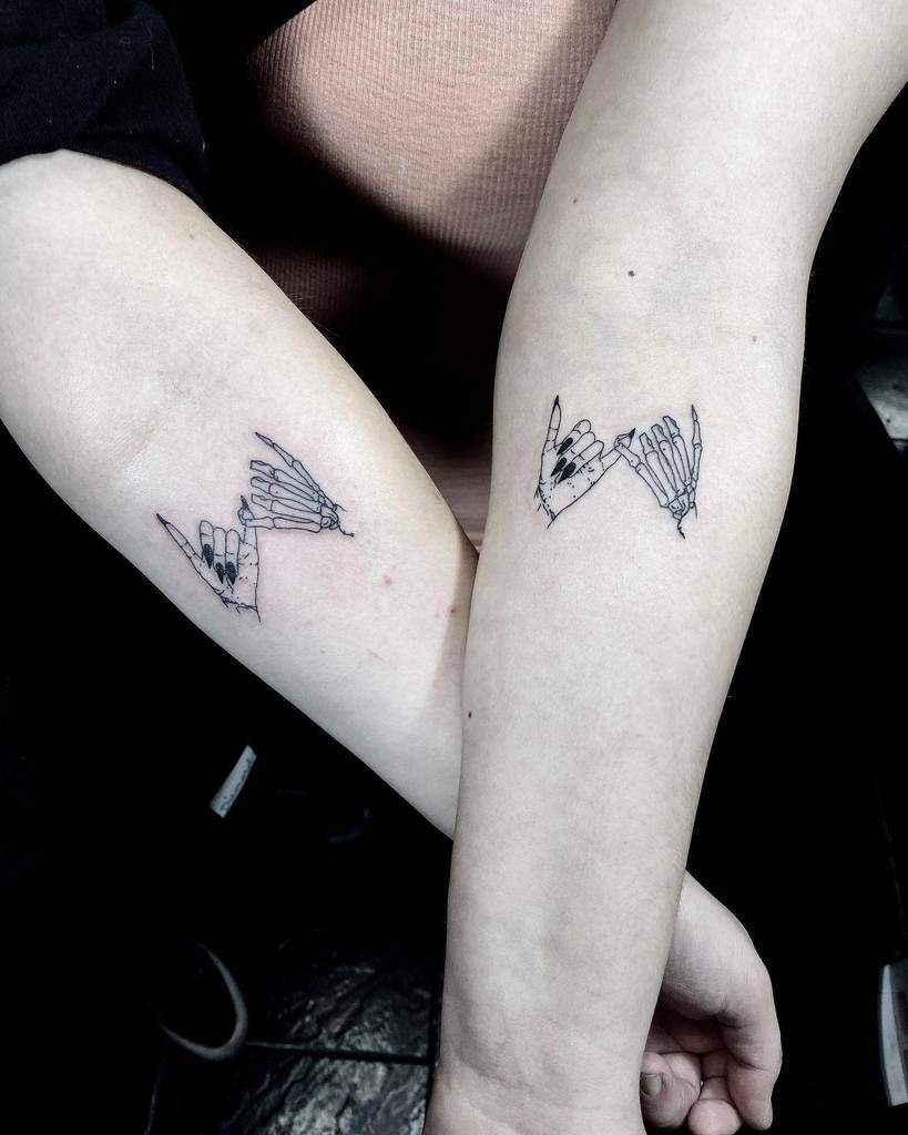 Matching pinky promise tattoos of a human hand and a skeleton hand with intricate details, placed on the inner forearms of two individuals