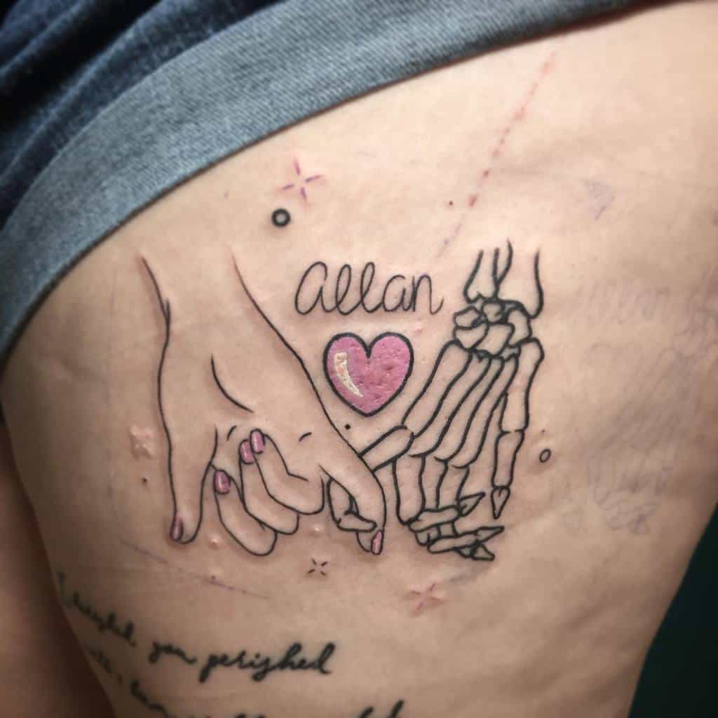 Pinky promise tattoo of a human and skeleton hand with a pink heart and the name "Allan," placed on the side of the torso