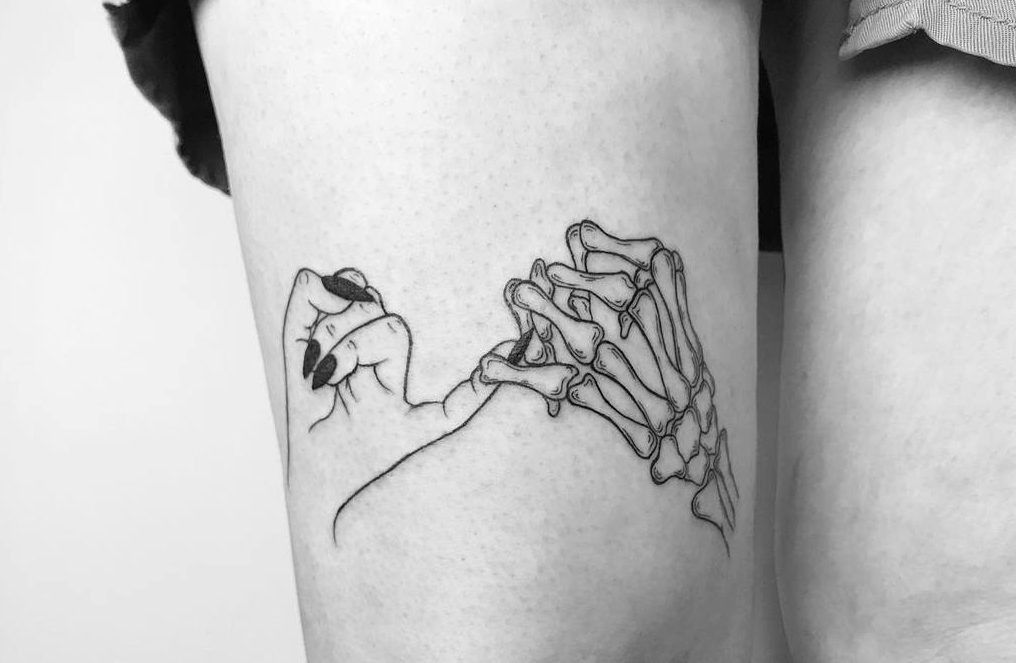 Black and white pinky promise tattoo featuring a human hand with black nails linked with a detailed skeleton hand, placed on the thigh