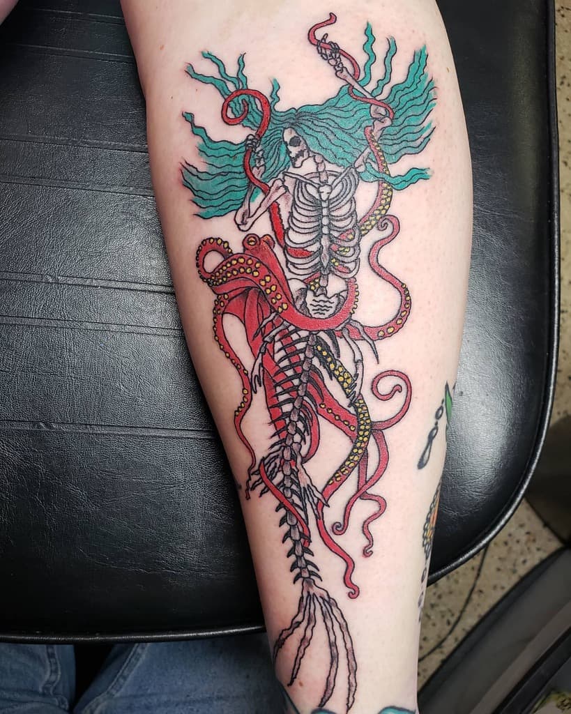 Colorful tattoo of a skeletal mermaid with flowing blue hair, wrapped in red octopus tentacles, blending dark fantasy and nautical themes.