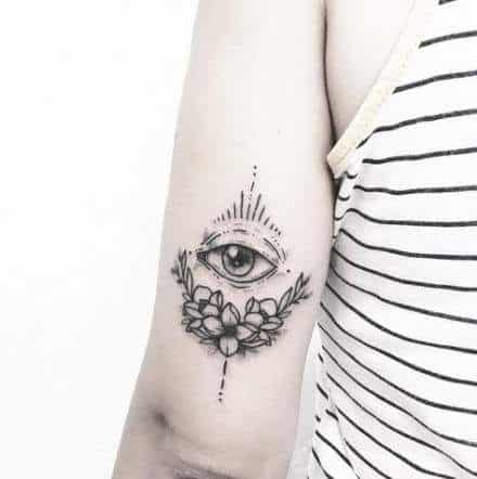Tattoo of an all-seeing eye with radiating lines, framed by blooming flowers, inked in fine black lines on the upper arm.