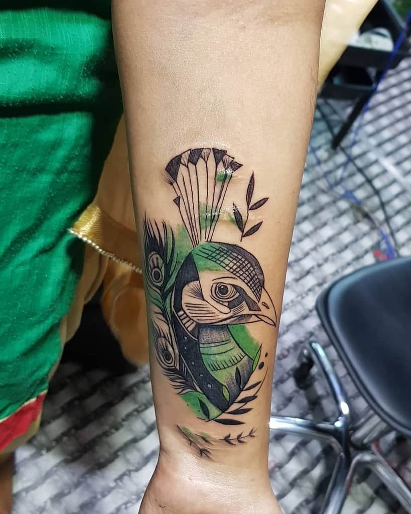 Geometric and abstract peacock tattoo on the forearm, featuring bold black ink with green shading, intricate patterns, and artistic detailing.