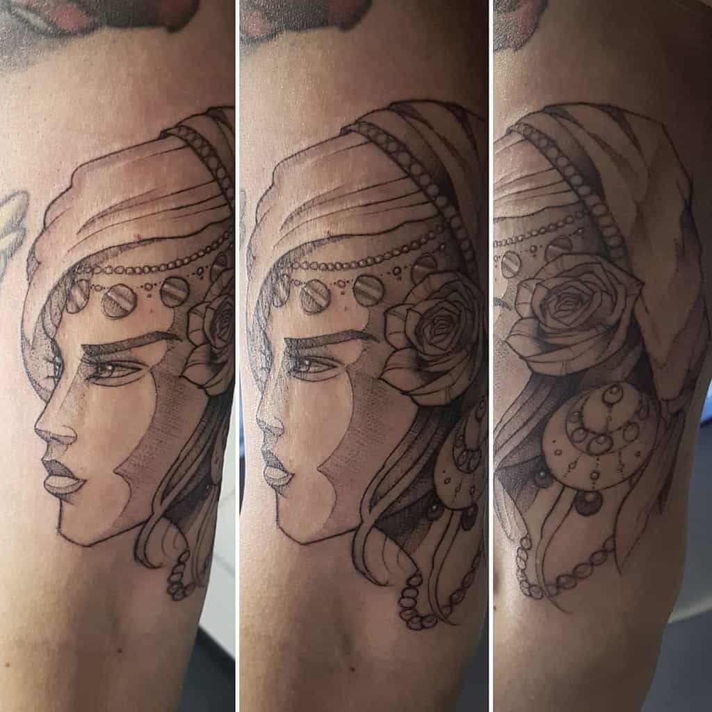 Black and grey tattoo of a gypsy woman in profile with a headscarf, beaded jewelry, a rose, and fine line shading on the arm.