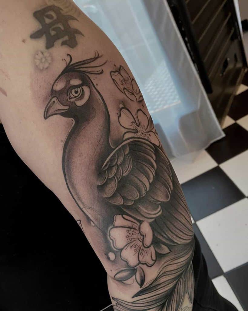 Black and grey peacock tattoo with soft shading, flowers, and detailed feathers on the upper arm.