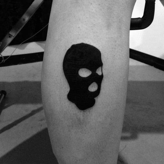 Minimalist tattoo of a solid black ski mask with open eye and mouth holes, placed on the back of the leg.