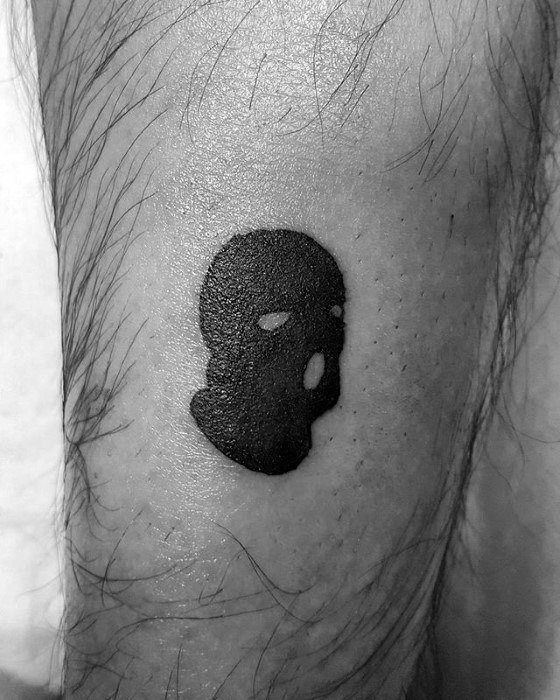 Small black ski mask tattoo with basic eye and mouth holes, inked on the lower leg with a bold silhouette style.
