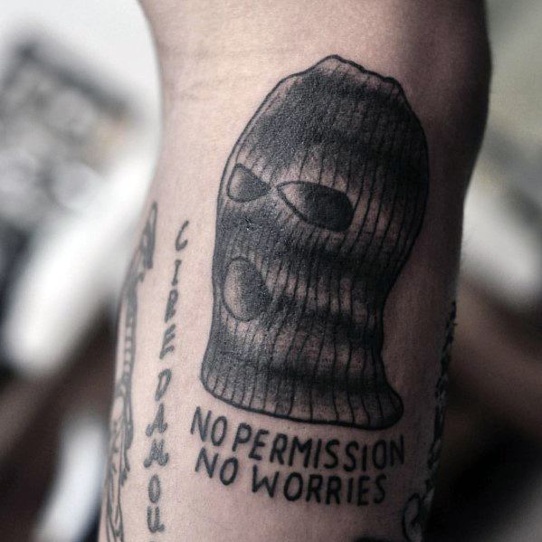 Tattoo of a shaded ski mask with the text NO PERMISSION NO WORRIES beneath it, inked in bold capital letters.