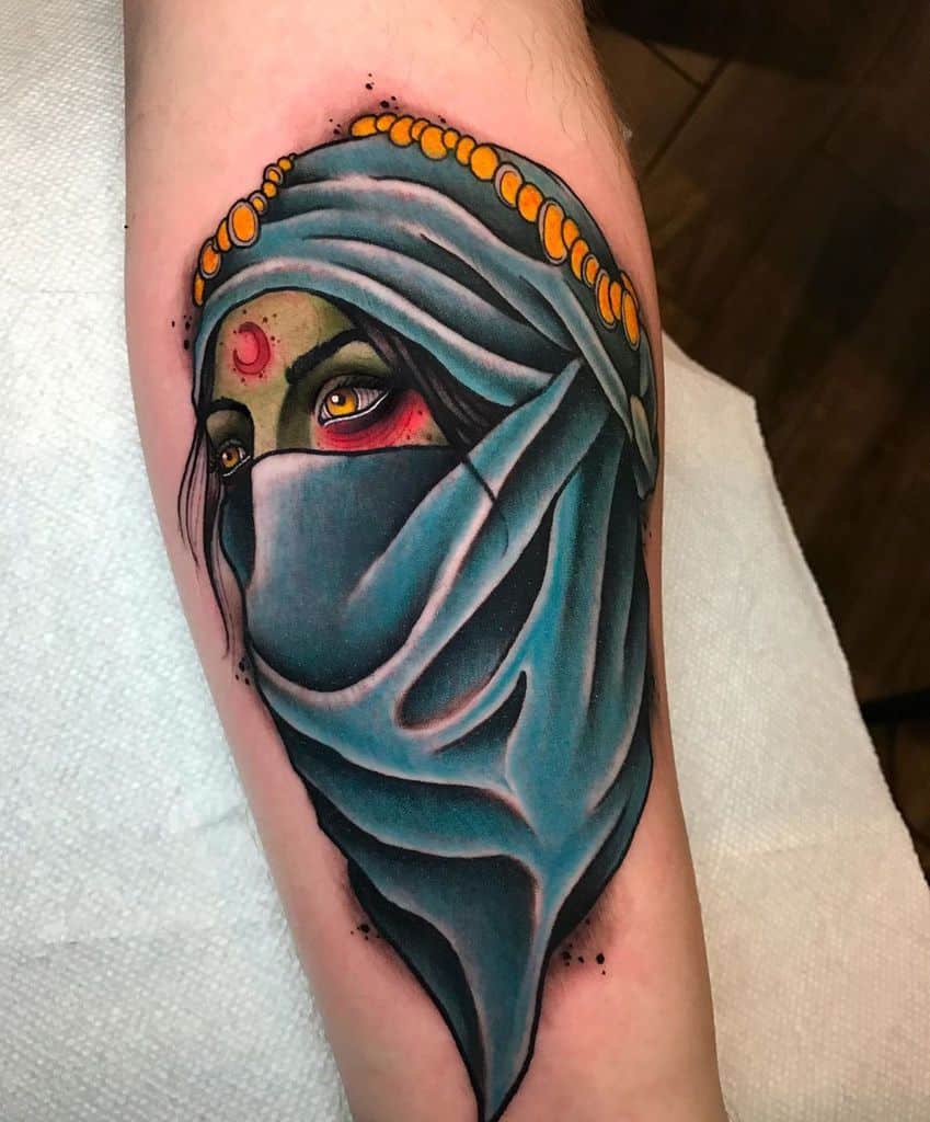 Vibrant tattoo of a mysterious veiled woman with piercing yellow eyes, a red forehead marking, and intricate headpiece details on the forearm.