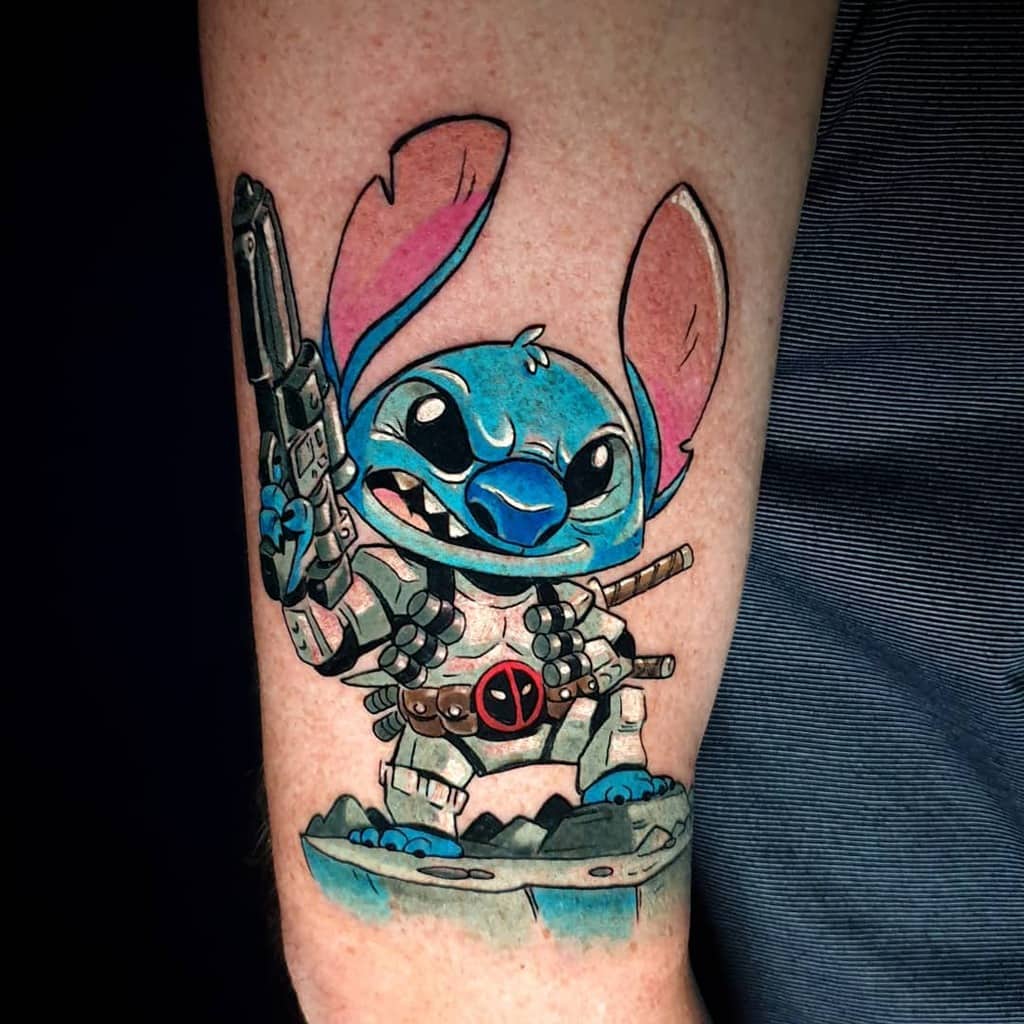 Colorful tattoo of Stitch from Lilo & Stitch dressed as Deadpool with weapons, on the forearm.
