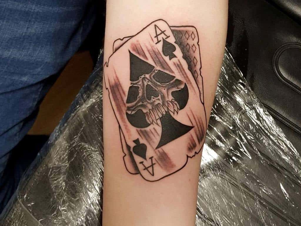 A black-and-white tattoo of the Ace of Spades card with a skull in the center, the card's edges torn
