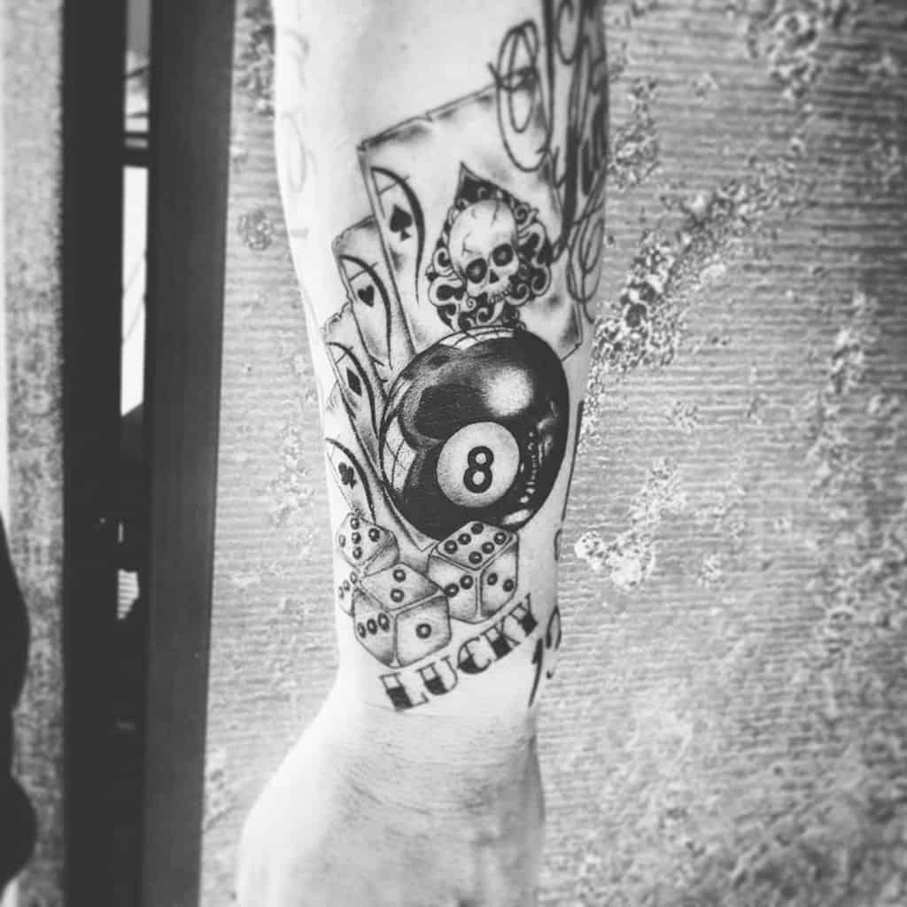 A black-and-white tattoo featuring the Ace of Spades cards, a skull, an 8-ball, dice, and the word "LUCKY" in a vintage style