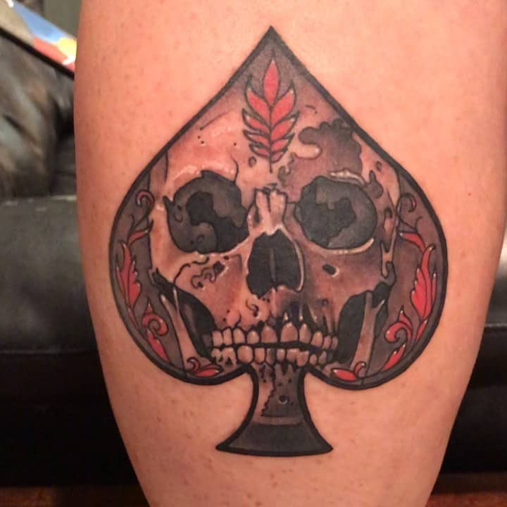 A tattoo featuring the Ace of Spades symbol, with a detailed skull in the center, surrounded by ornate red floral designs