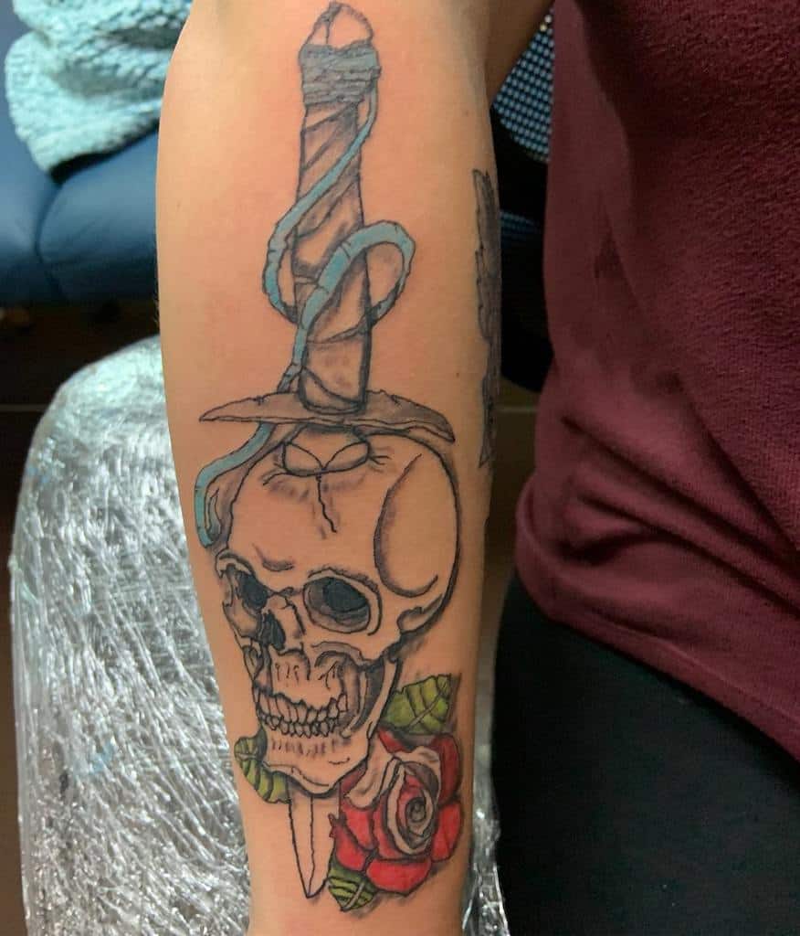Forearm tattoo of a skull impaled by a dagger wrapped in blue ribbon, with a red rose at the base, blending dark and traditional elements.