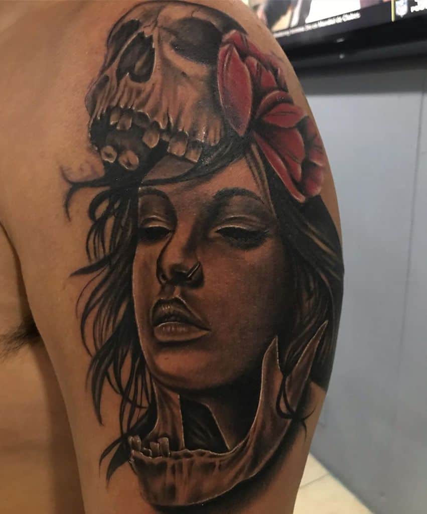 A highly detailed tattoo on the upper arm featuring a realistic woman's face, a skull, a red flower, and a broken jawbone for a dramatic effect.