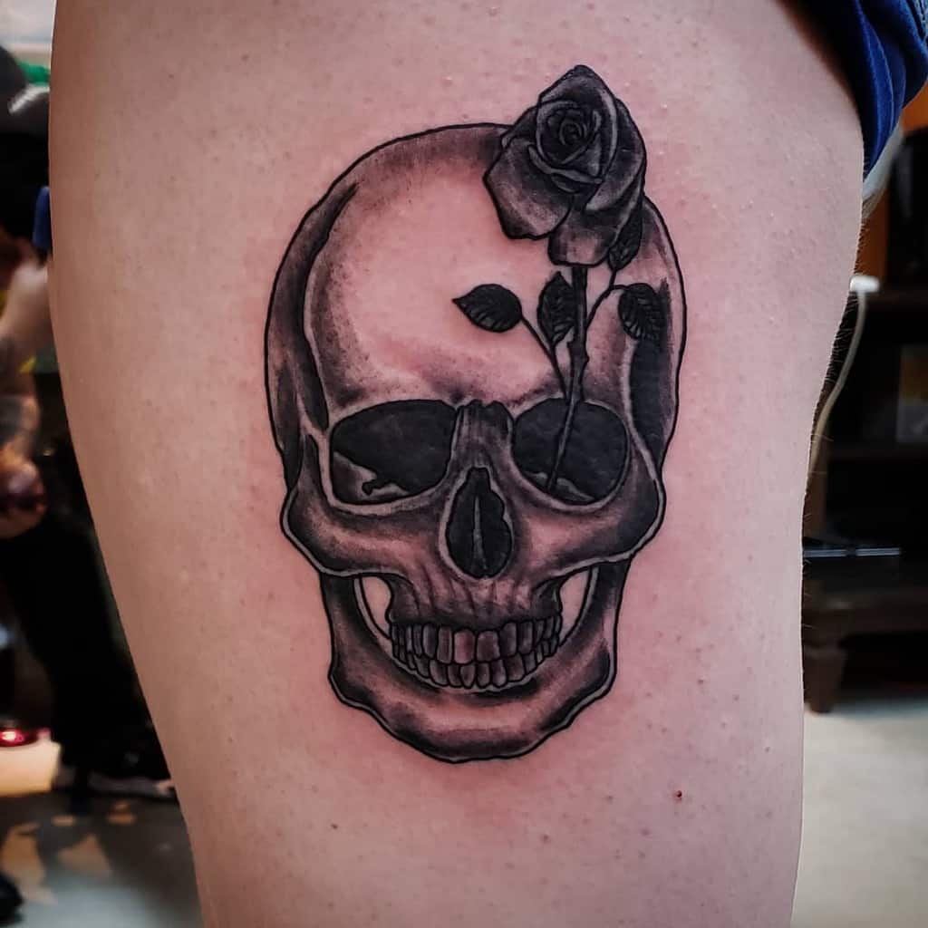 Black and grey thigh tattoo of a skull with a delicate rose growing from its head, blending dark symbolism with elegance.