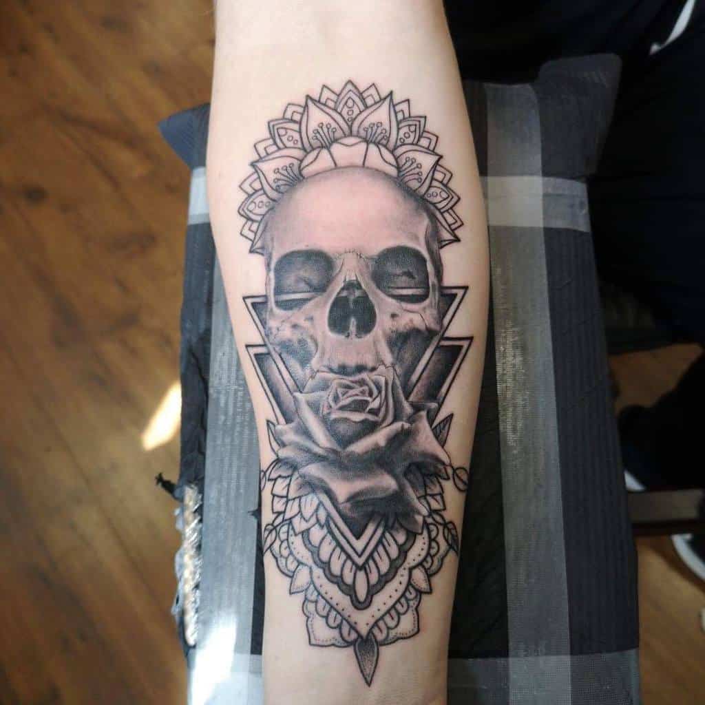 Intricate black and grey tattoo of a skull with a rose, geometric shapes, and mandala patterns, creating a detailed forearm design.
