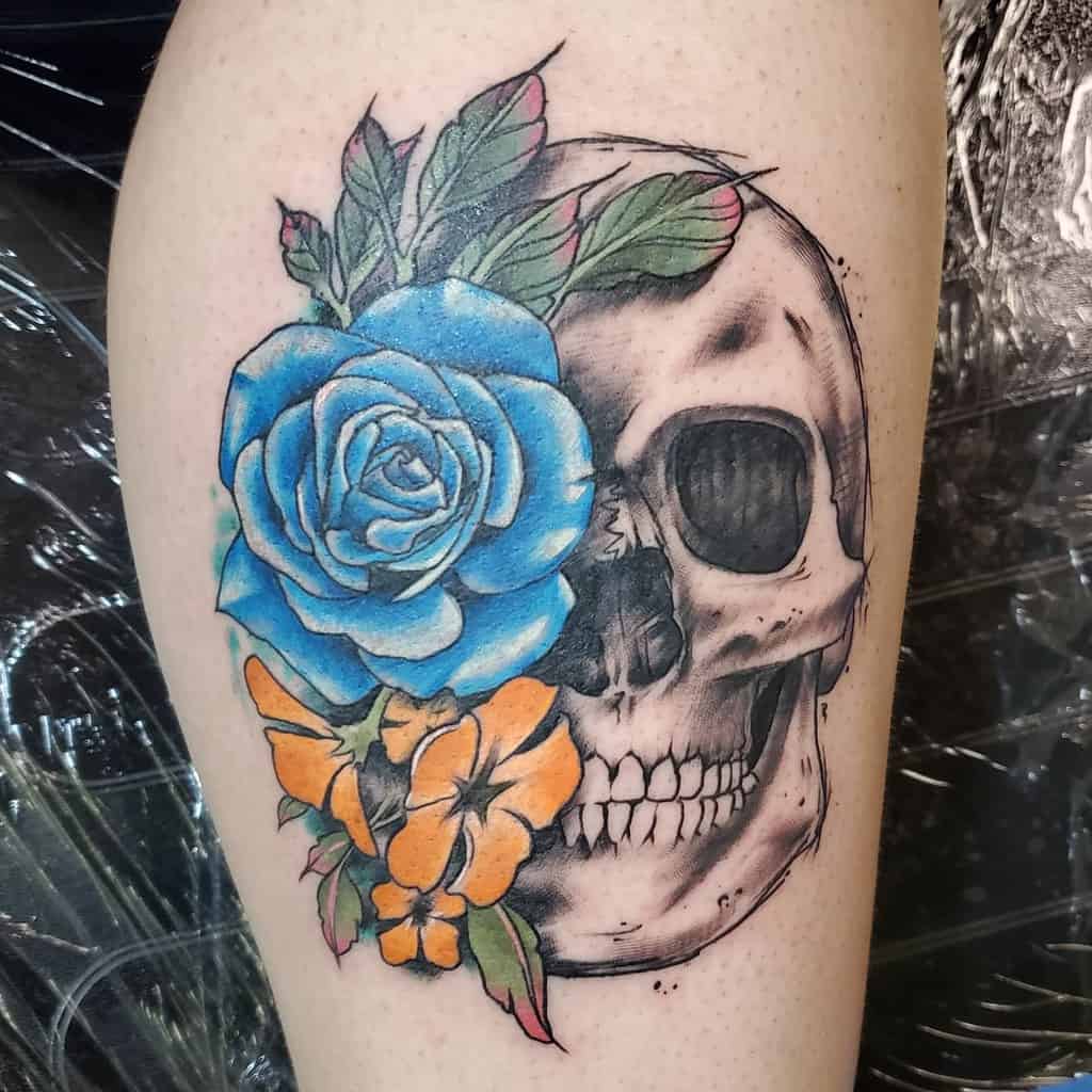 Vibrant thigh tattoo of a skull adorned with a blue rose and orange flowers, blending realism with bold color contrasts.