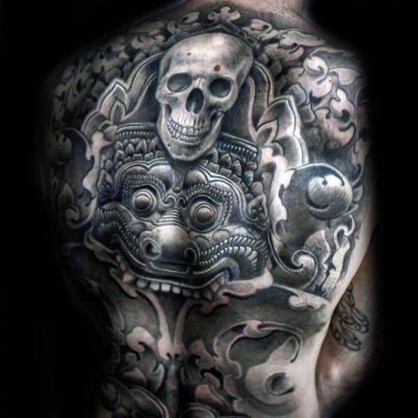 Ornate back tattoo featuring a detailed skull above a traditional Balinese-style demon mask, framed by swirling smoke and patterns.