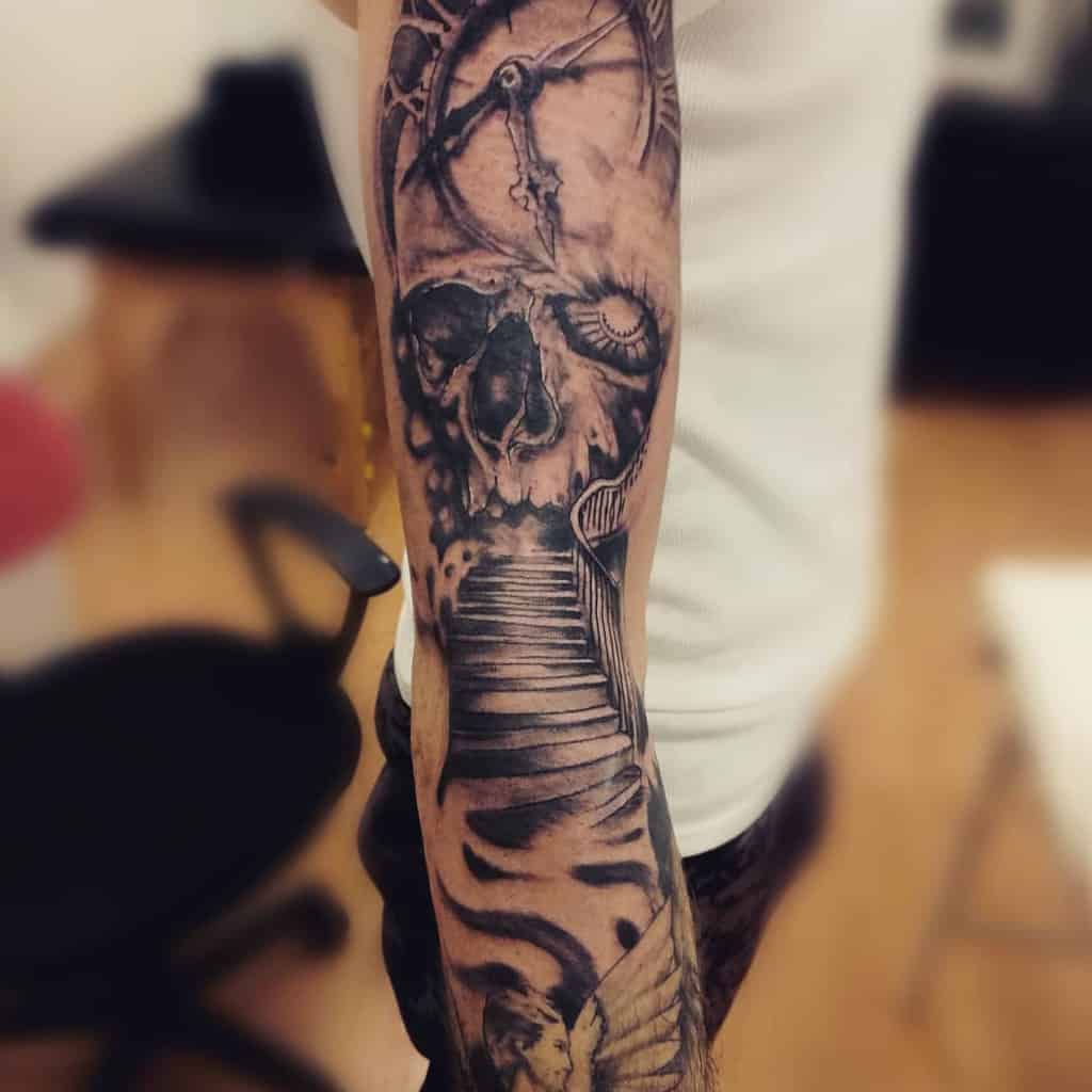Black and grey full sleeve tattoo featuring a detailed skull, clock, and staircase, creating a haunting and dramatic design