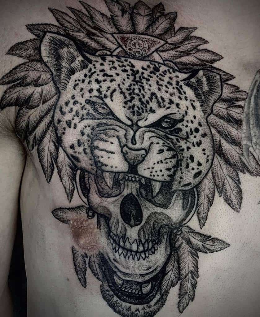 A striking tattoo of a jaguar's face with intense details, surrounded by leaf patterns and a skull at the bottom, with a symbolic third eye design at the top, placed on the chest.