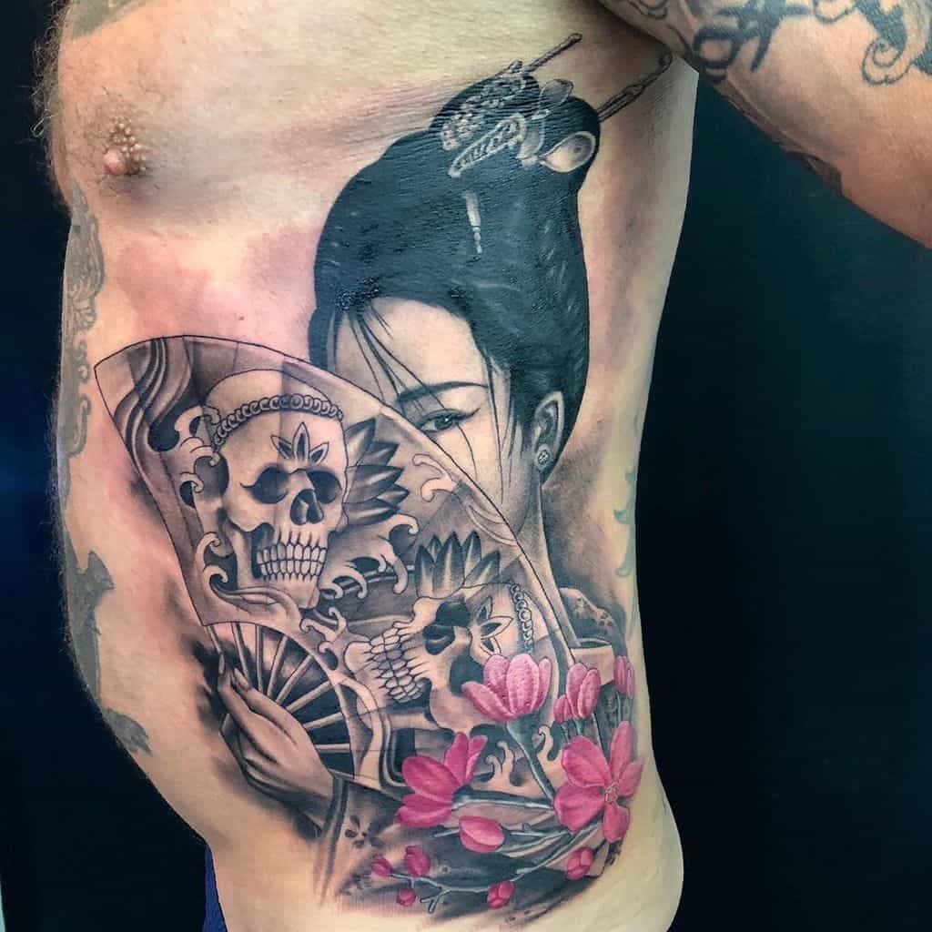 Black and grey rib tattoo of a Geisha holding a fan decorated with skulls, accented with pink cherry blossoms for a striking contrast.