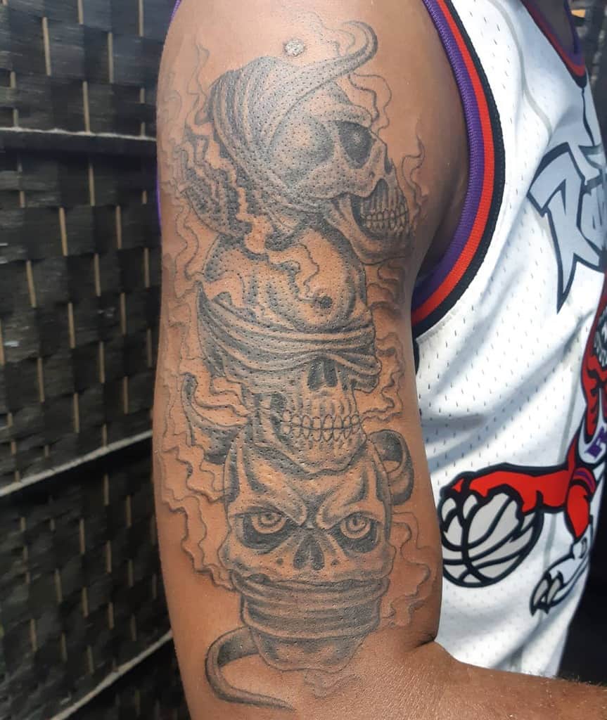 Detailed black and gray tattoo of three skulls on an upper arm, representing 'Hear No Evil, See No Evil, Speak No Evil,' with smoky effects and bold shading