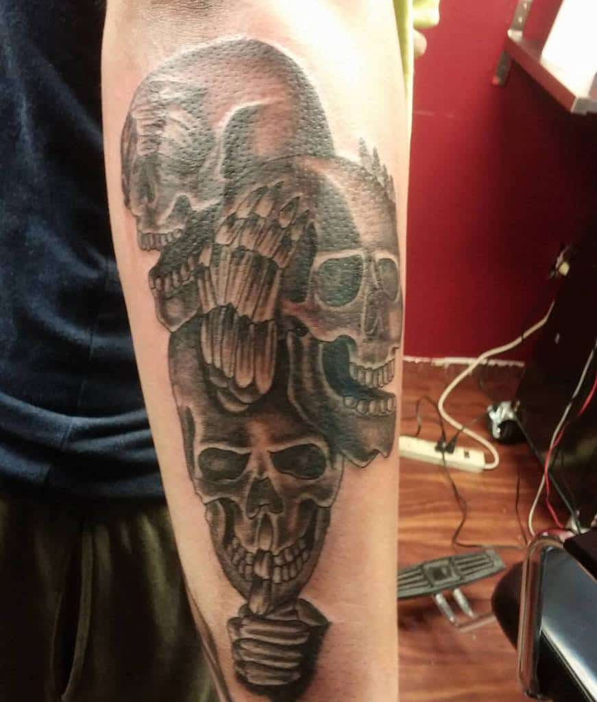 Dark, shaded tattoo of three skulls stacked vertically on a forearm, symbolizing 'Hear No Evil, See No Evil, Speak No Evil,' with skeletal hands covering ears, eyes, and mouth