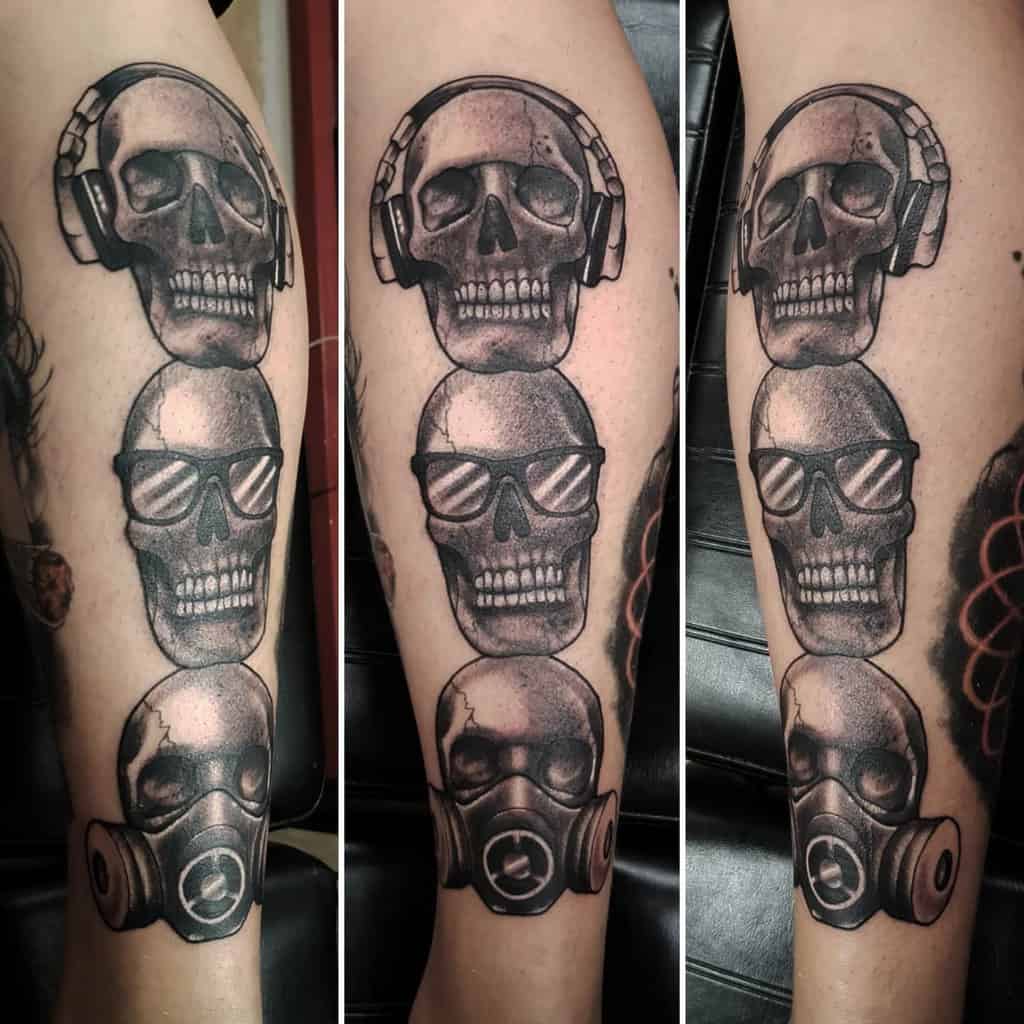 Three skull tattoos in a vertical line, wearing headphones, sunglasses, and a gas mask on a leg