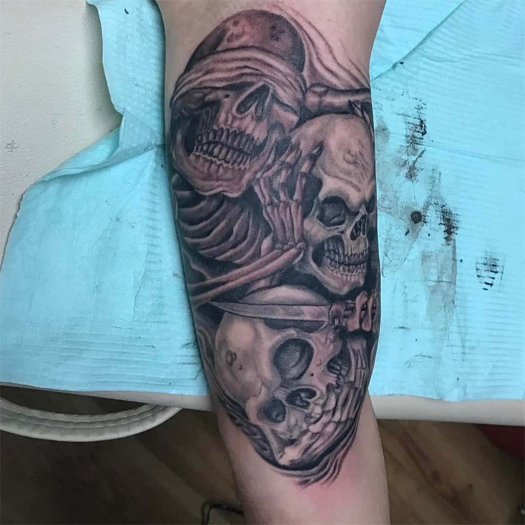 Detailed tattoo of three skulls representing 'Hear No Evil, See No Evil, Speak No Evil,' with blindfolds and skeletal hands in shaded black and gray tones