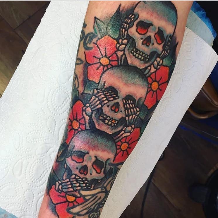 Colorful tattoo of three skulls representing 'Hear No Evil, See No Evil, Speak No Evil,' surrounded by vibrant flowers