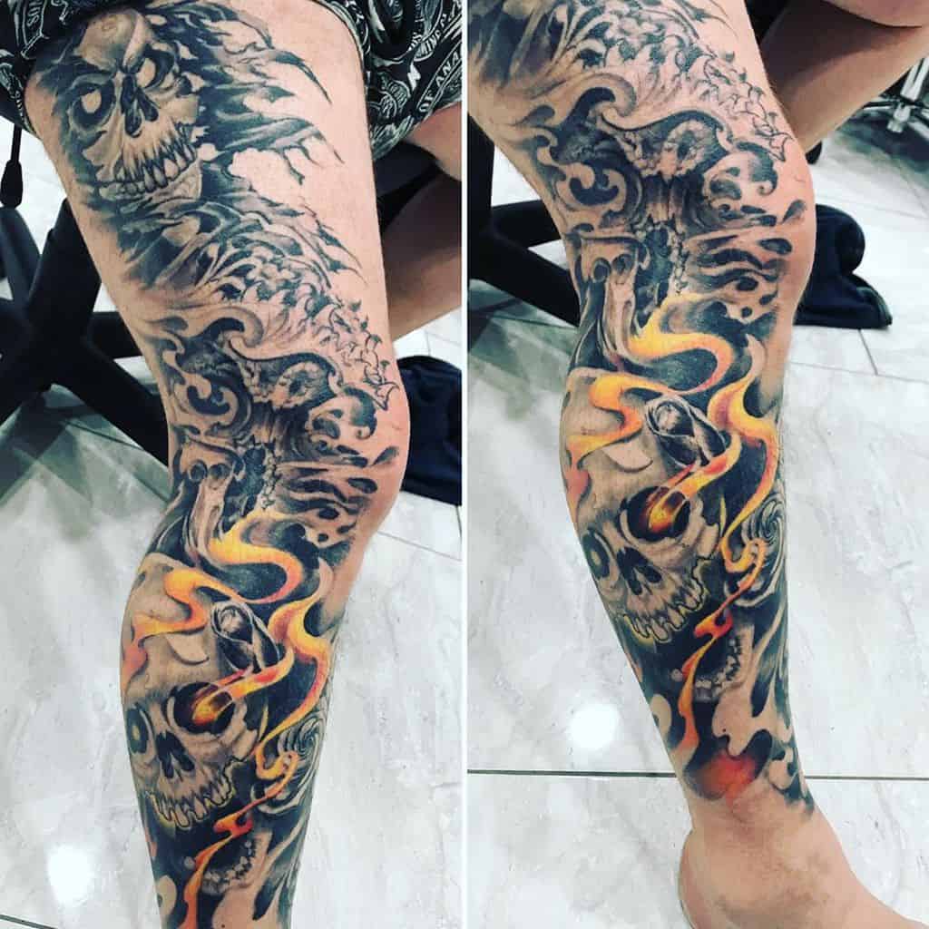 A leg sleeve tattoo featuring a skull with flames and swirling clouds in black and grey tones, accentuated with orange and yellow
