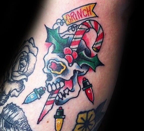 Christmas tattoo of a skull decorated with holly, a candy cane, and colorful holiday lights, featuring a "Grinch" banner for a festive yet edgy design.