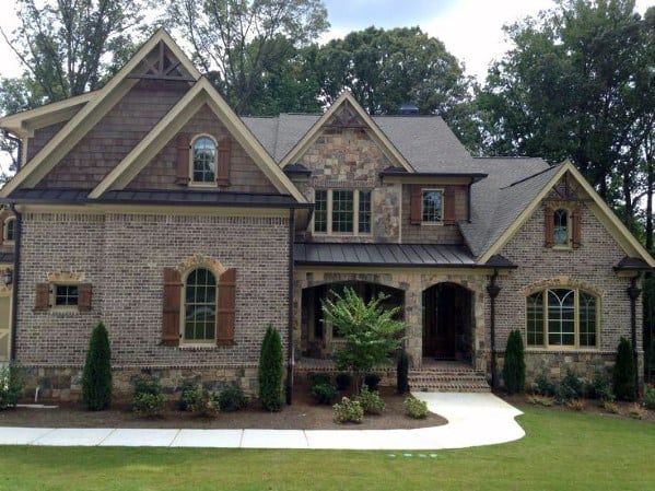 Charming home with rustic brick and stone cladding, wooden shutters, gabled rooflines, and a welcoming porch surrounded by greenery