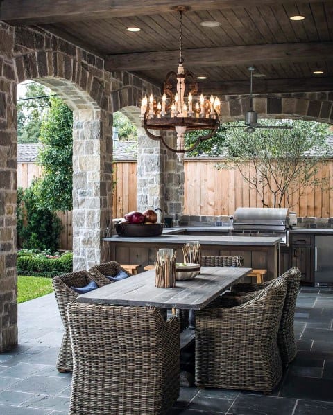 Outdoor patio featuring wicker chairs, a wooden dining table, stone arches, a chandelier, and a built-in grill